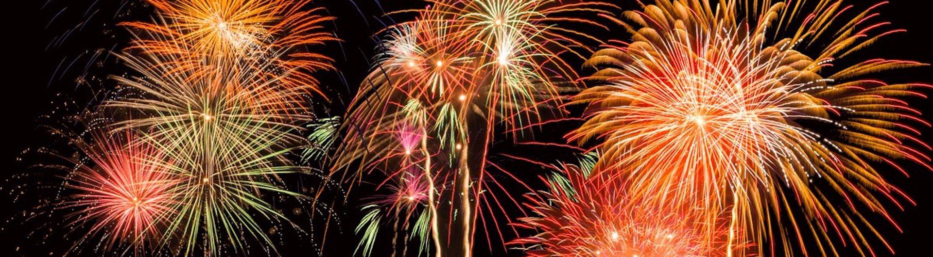 Colorful fireworks burst in the night sky, displaying vibrant red, orange, yellow, and green patterns against a black background. Several fireworks are exploding simultaneously, creating a festive atmosphere.