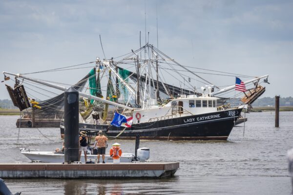 A fishing boat named LADY CATHERINE with an American flag docked in a harbor; people stand on a small boat nearby, and other fishing boats are visible in the background.