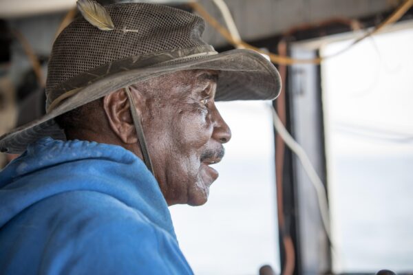 An elderly man wearing a wide-brimmed hat with a feather and a blue hoodie looks thoughtfully out a window, with soft light illuminating his face.