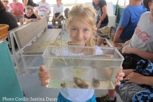 A smiling young girl on a boat holds a clear plastic container filled with water, small marine creatures, and a piece of coral, surrounded by seated people. Photo Credit: Justina Dacey.
