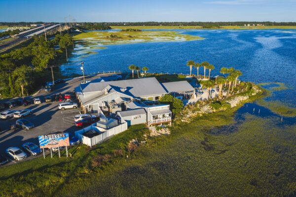 Aerial view of Shuckers restaurant on Amelia Island, surrounded by blue water and marshland with a bridge in the background and cars parked nearby under a clear sky.