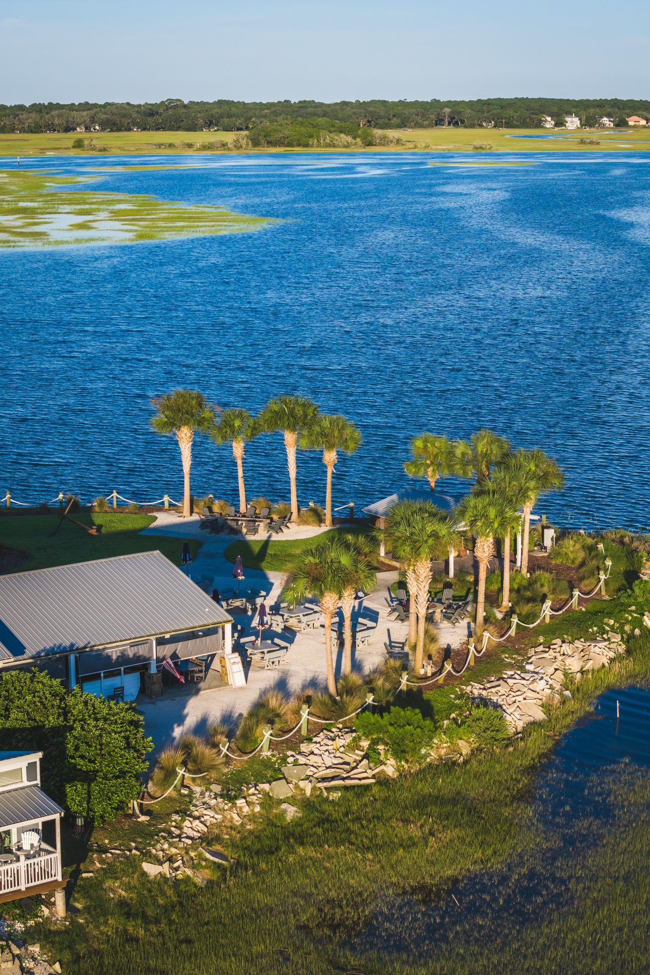 Aerial view of a lakeside area with palm trees, a small building, outdoor seating, and a stone-bordered shoreline, surrounded by blue water and grassy patches. Houses and trees are visible in the distant background.