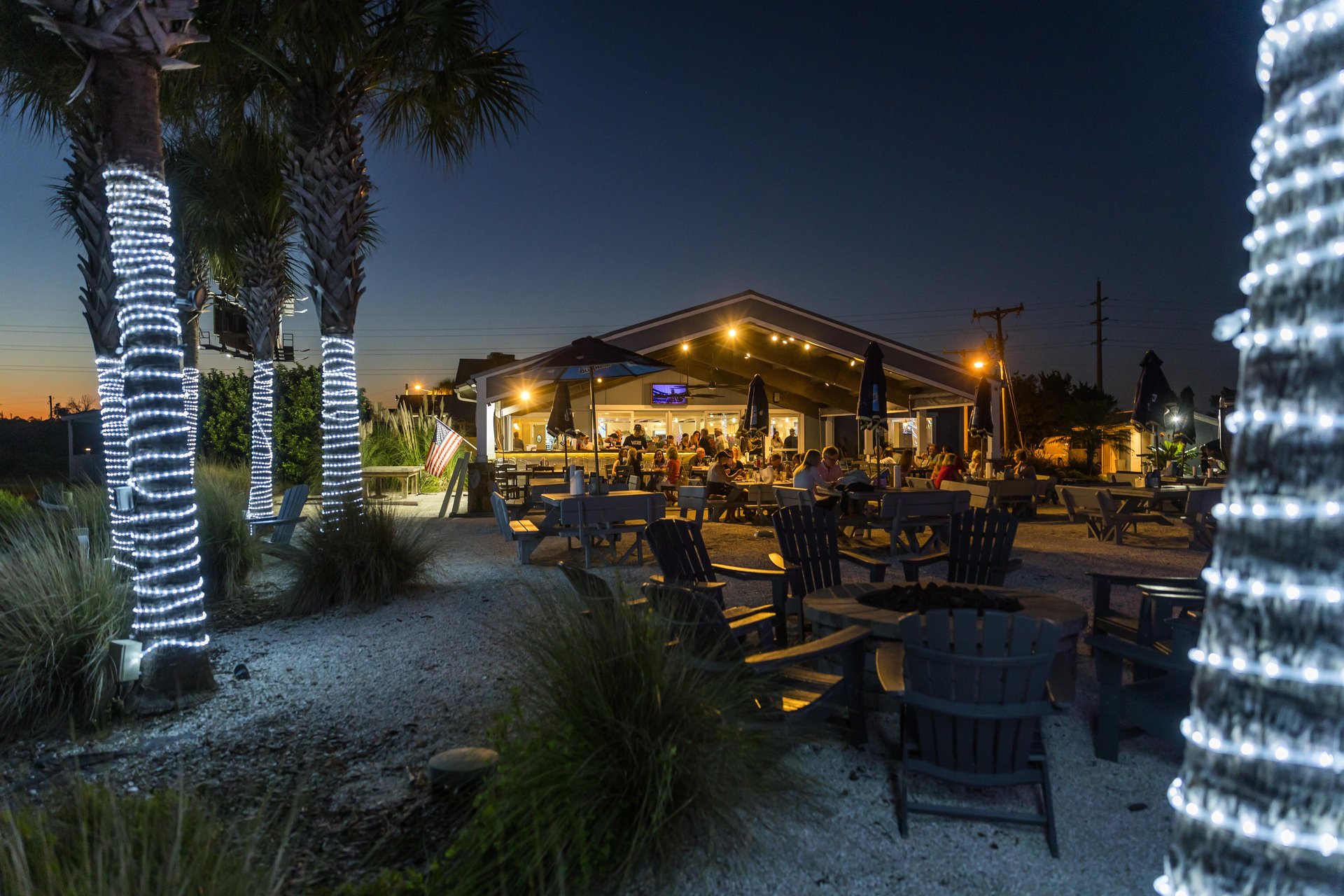 An outdoor restaurant at dusk with people dining, surrounded by palm trees wrapped in white string lights. Adirondack chairs and tables are set on a gravel surface under a darkening sky.
