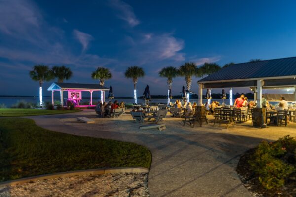 An outdoor restaurant by the water at dusk features people dining at tables, palm trees wrapped in lights, a stage with purple lighting, and a relaxed, festive atmosphere.