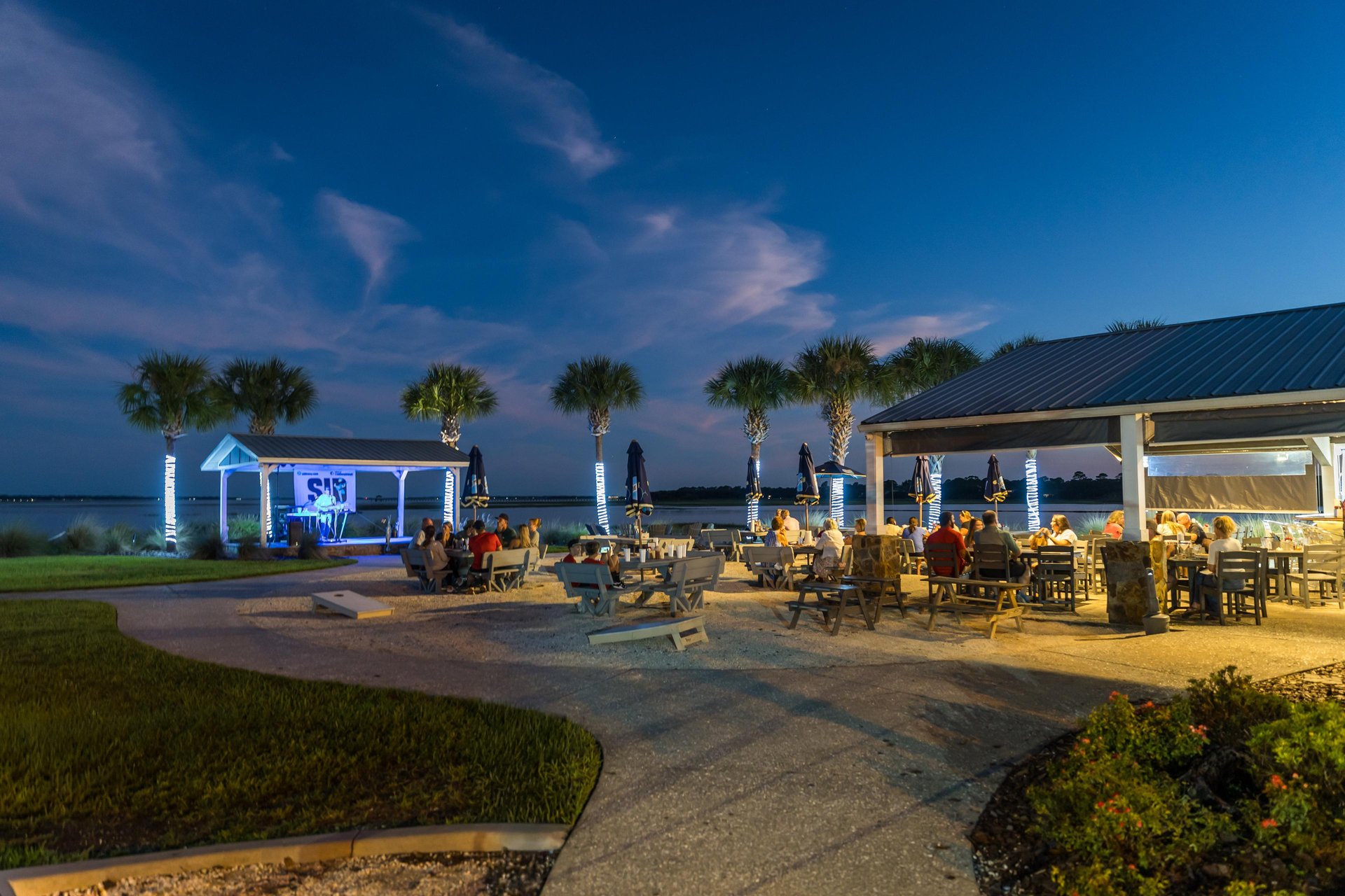An outdoor restaurant with people dining under a pavilion at sunset, surrounded by palm trees and overlooking the water, with a small stage featuring live music in the background.