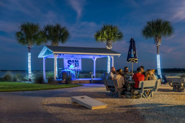 A group of people sit at picnic tables near the water at Shuckers on Amelia Island during sunset. A musician performs under a blue-lit gazebo surrounded by palm trees wrapped in string lights, with a cornhole game set up nearby.