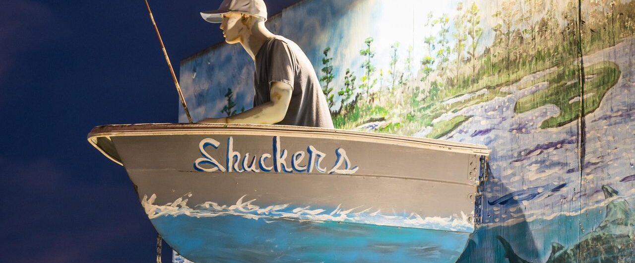 A boat labeled “Shuckers” with a mannequin fisherman is mounted on a mural-covered wall, illuminated by a spotlight at night, with a twilight sky in the background.