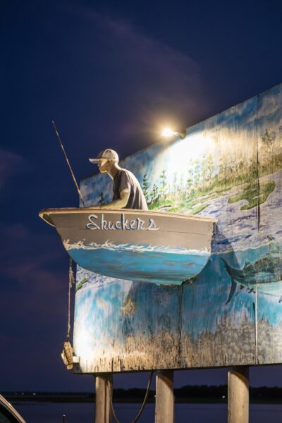 A boat labeled “Shuckers” with a mannequin fisherman is mounted on a mural-covered wall, illuminated by a spotlight at night, with a twilight sky in the background.