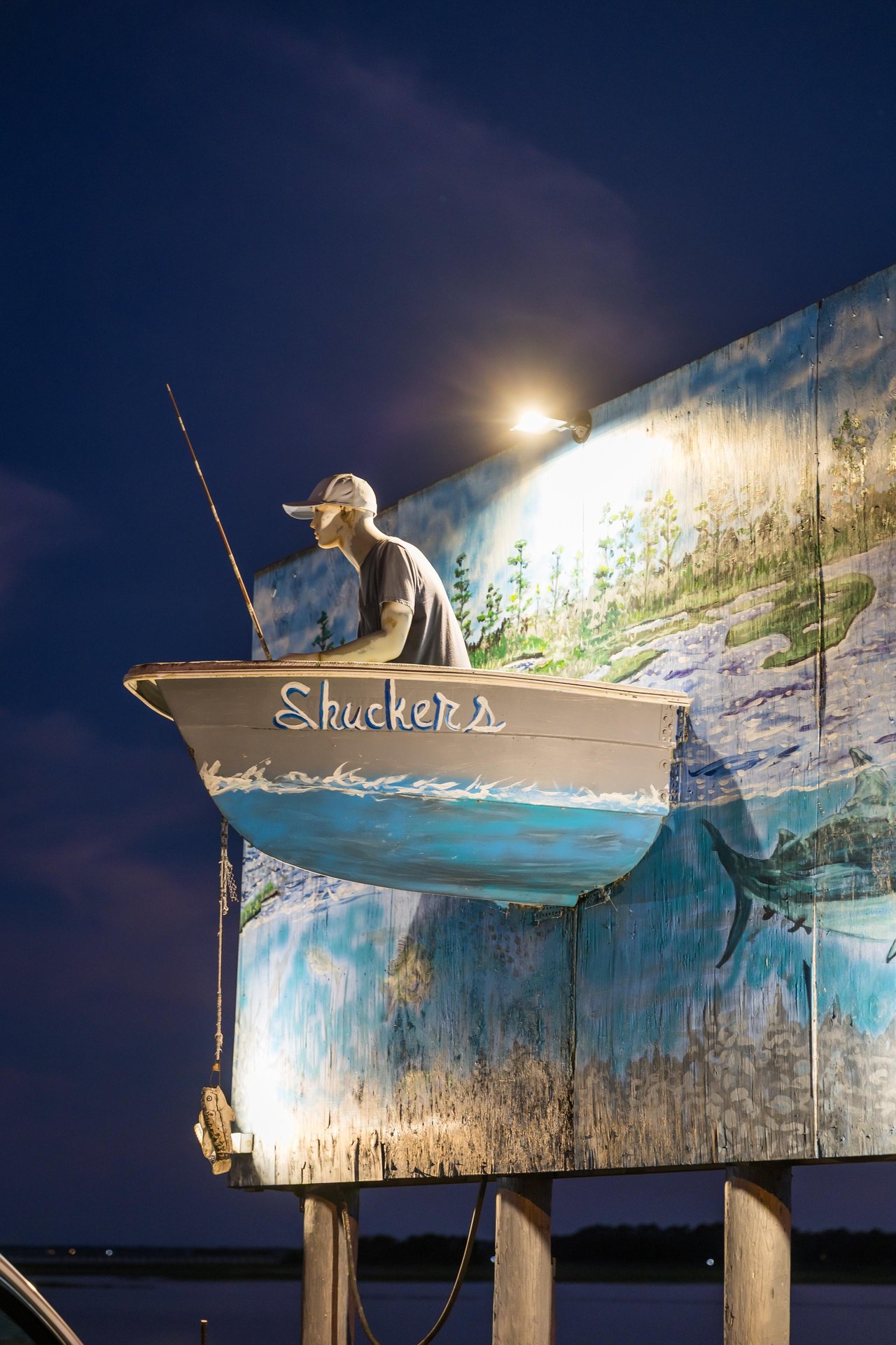 A boat labeled “Shuckers” with a mannequin fisherman is mounted on a mural-covered wall, illuminated by a spotlight at night, with a twilight sky in the background.