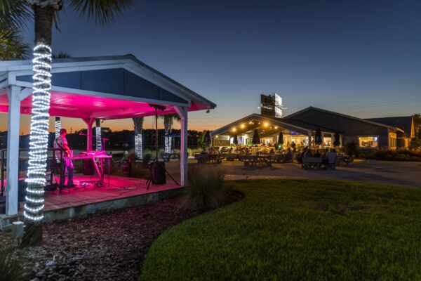 At sunset, a musician performs on an outdoor stage illuminated with pink lighting, while guests dine at Shuckers on Amelia Island. String lights and palm trees surround the open-air restaurant, creating a lively coastal evening atmosphere.