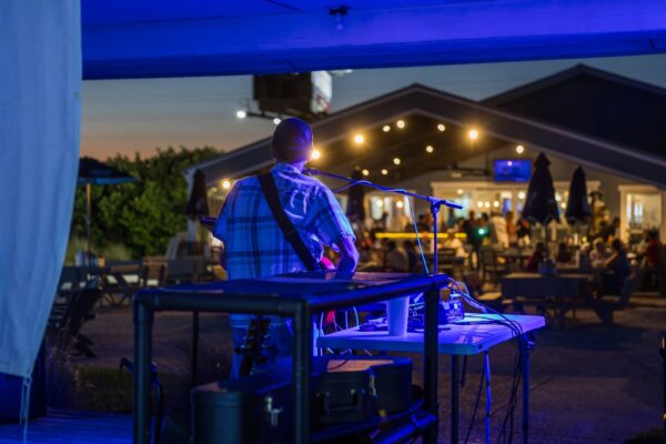 A musician performs at Shuckers on Amelia Island in the evening, playing guitar and singing into a microphone under blue stage lights. The view looks toward the outdoor dining area, where guests sit beneath string lights glowing warmly against the night sky.