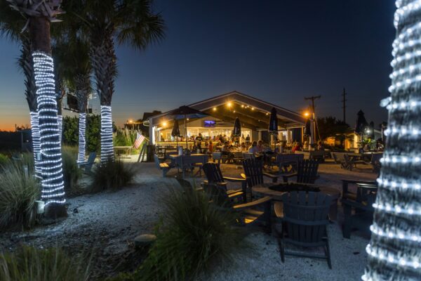 An outdoor dining area at Shuckers on Amelia Island at dusk, with people enjoying food and drinks under a glowing pavilion. Palm trees wrapped in white string lights surround Adirondack chairs and tables on a sandy patio, creating a cozy coastal atmosphere.