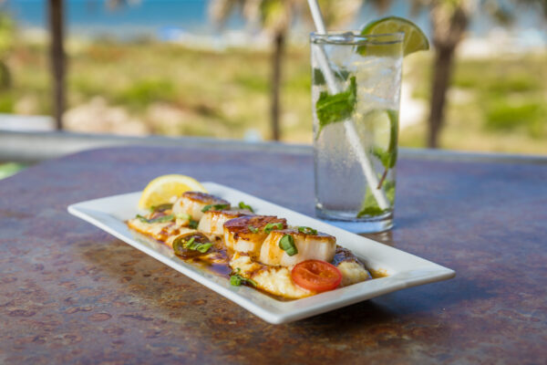 A rectangular plate of grilled scallops garnished with herbs, a lemon wedge, and cherry tomato sits on a table next to a refreshing drink with lime, mint, and ice. Palm trees and a beach are blurred in the background.