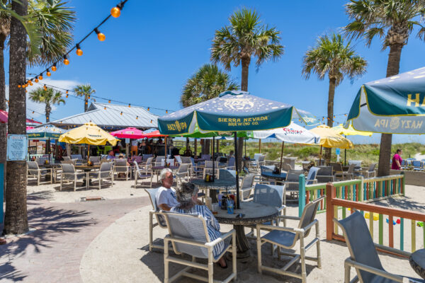 People sit at outdoor tables under colorful umbrellas at a beachside restaurant surrounded by palm trees on a sunny day. String lights hang overhead, and a sandy area and grassy dunes are visible in the background.