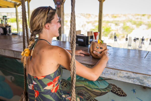 A woman in a floral dress sits on a swing at an outdoor bar, holding a coconut drink with a straw. The bar counter has bottles and napkins, and the background shows a sunny, sandy landscape.