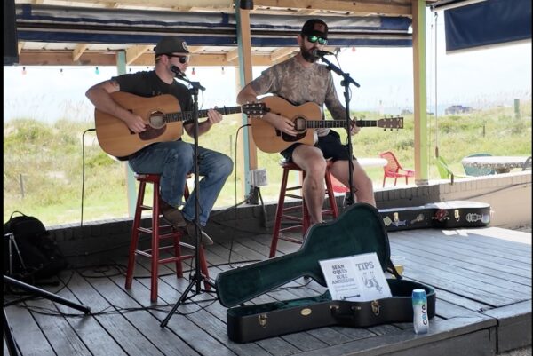 Two musicians perform at Sliders Seaside Grill, sitting on stools with acoustic guitars and microphones on an outdoor stage overlooking the dunes. A guitar case with a tips sign rests in front of them.