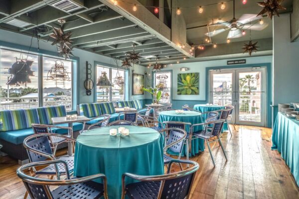 A bright, airy room with teal tablecloths, metal chairs, and large windows. String lights and star-shaped lamps hang from the ceiling, and sunlight streams in, creating a relaxing, coastal atmosphere.