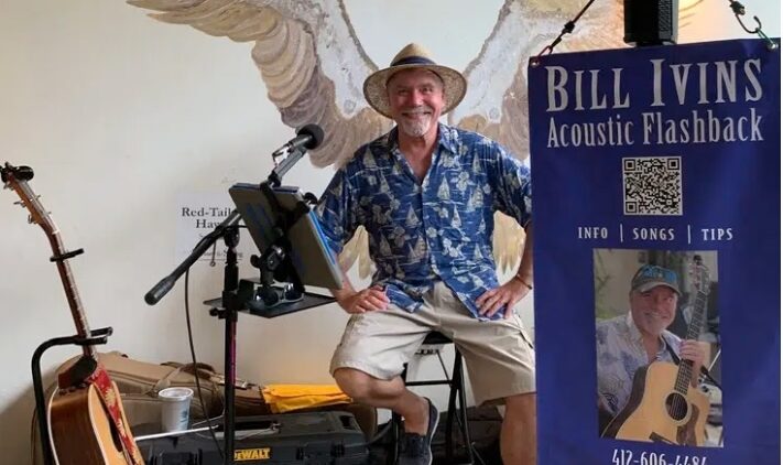 A smiling man in a straw hat and blue shirt sits on a stool with musical equipment and guitars, with painted wings on the wall behind him. A banner reads Bill Ivins Acoustic Flashback with contact info and a photo.