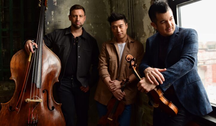 Three men stand indoors by a window, holding string instruments—a double bass, a viola, and a violin. They pose casually against a textured wall with soft natural light shining in.