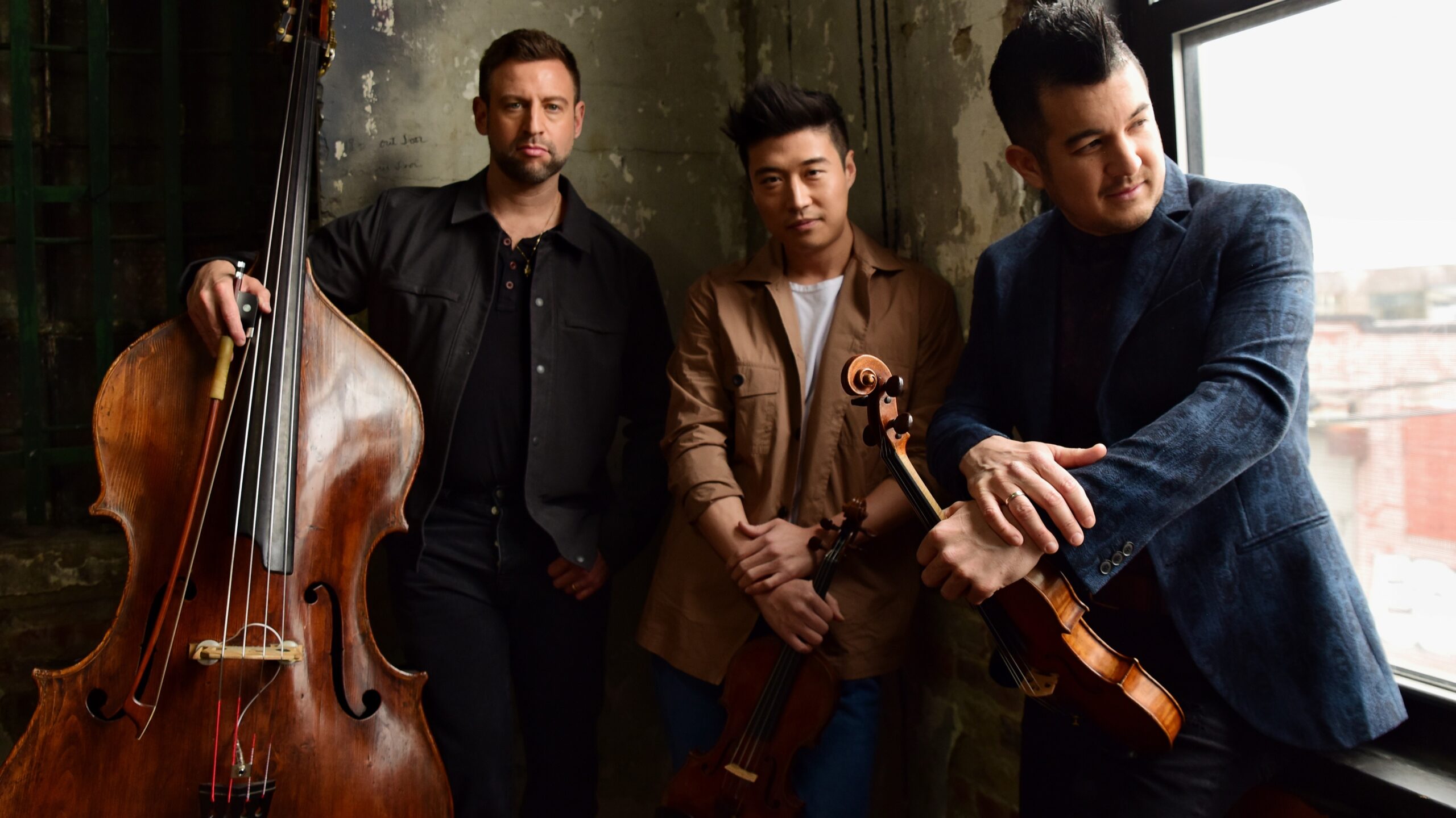 Three men stand indoors by a window, holding string instruments—a double bass, a viola, and a violin. They pose casually against a textured wall with soft natural light shining in.