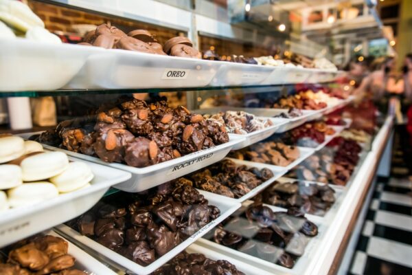 A display case filled with various types of chocolate candies and confections on white trays, labeled with names like “Oreo” and “Rocky Road,” in a brightly lit candy shop.