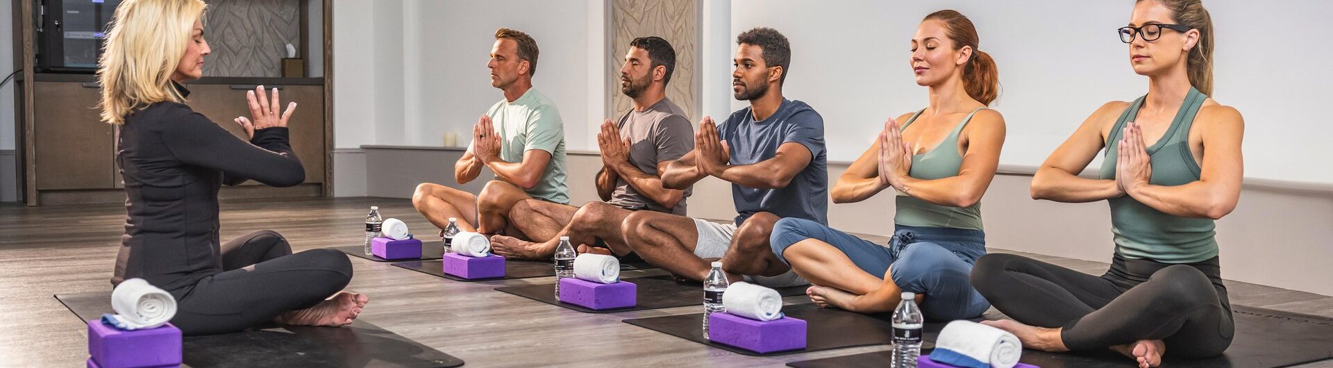 A yoga instructor leads five adults seated on mats in a studio, all in a cross-legged position with hands in prayer pose, facing the instructor. Yoga props and water bottles are placed beside them.