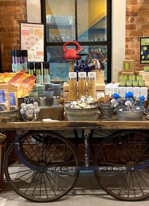 A rustic shop display with shelves of tea jars, a central cart holding tea accessories, tins, mugs, tea canisters, a red kettle, and colorful packaging; set against an exposed brick wall.