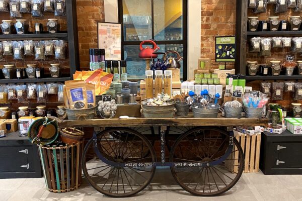 A rustic shop display with shelves of tea jars, a central cart holding tea accessories, tins, mugs, tea canisters, a red kettle, and colorful packaging; set against an exposed brick wall.
