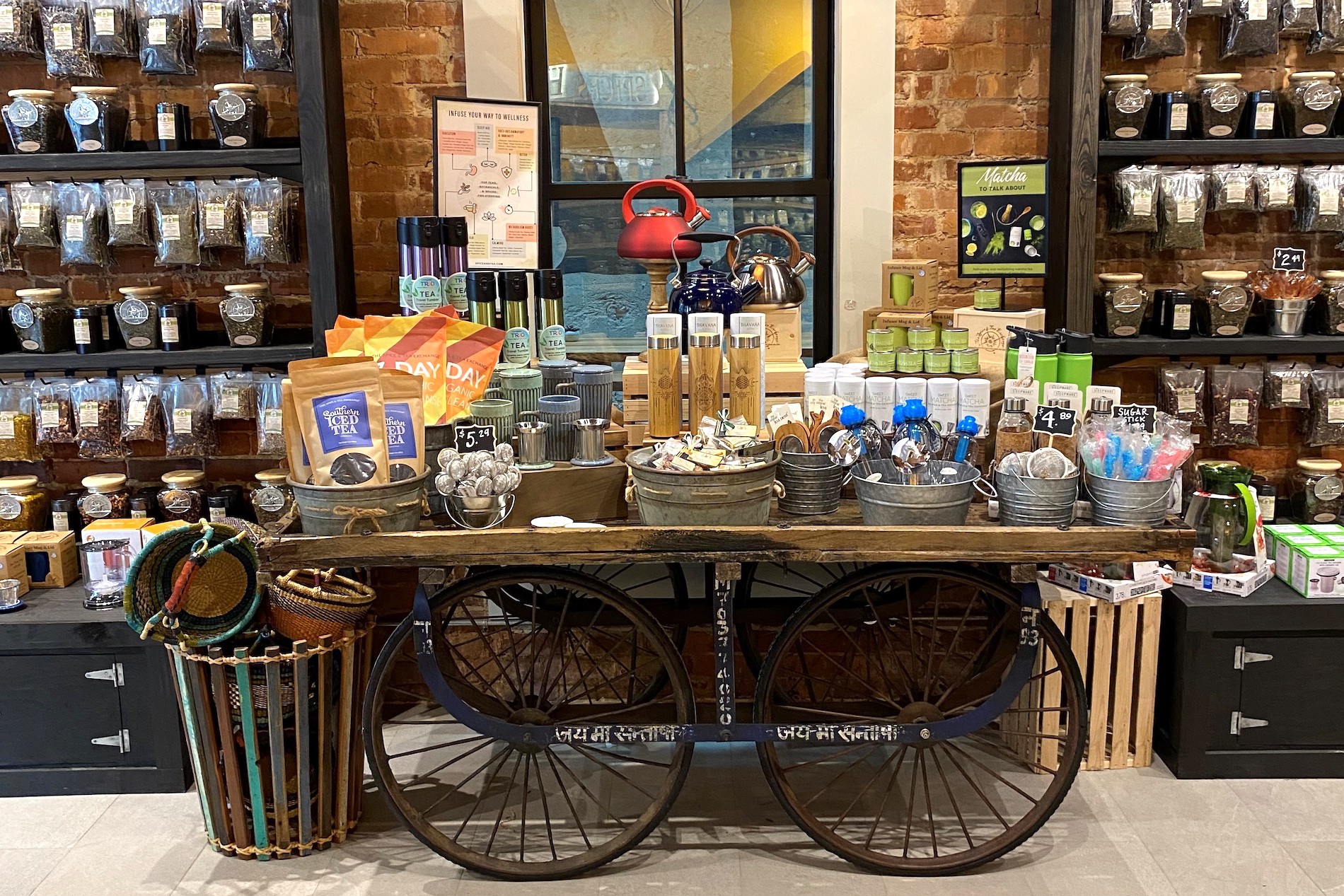 A rustic shop display with shelves of tea jars, a central cart holding tea accessories, tins, mugs, tea canisters, a red kettle, and colorful packaging; set against an exposed brick wall.