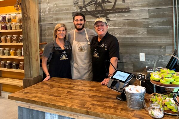 Three people stand smiling behind a wooden counter in a rustic store with shelves of jars and goods. Two wear black shirts with name tags, and one wears a light apron. A tablet register and small products are on the counter.