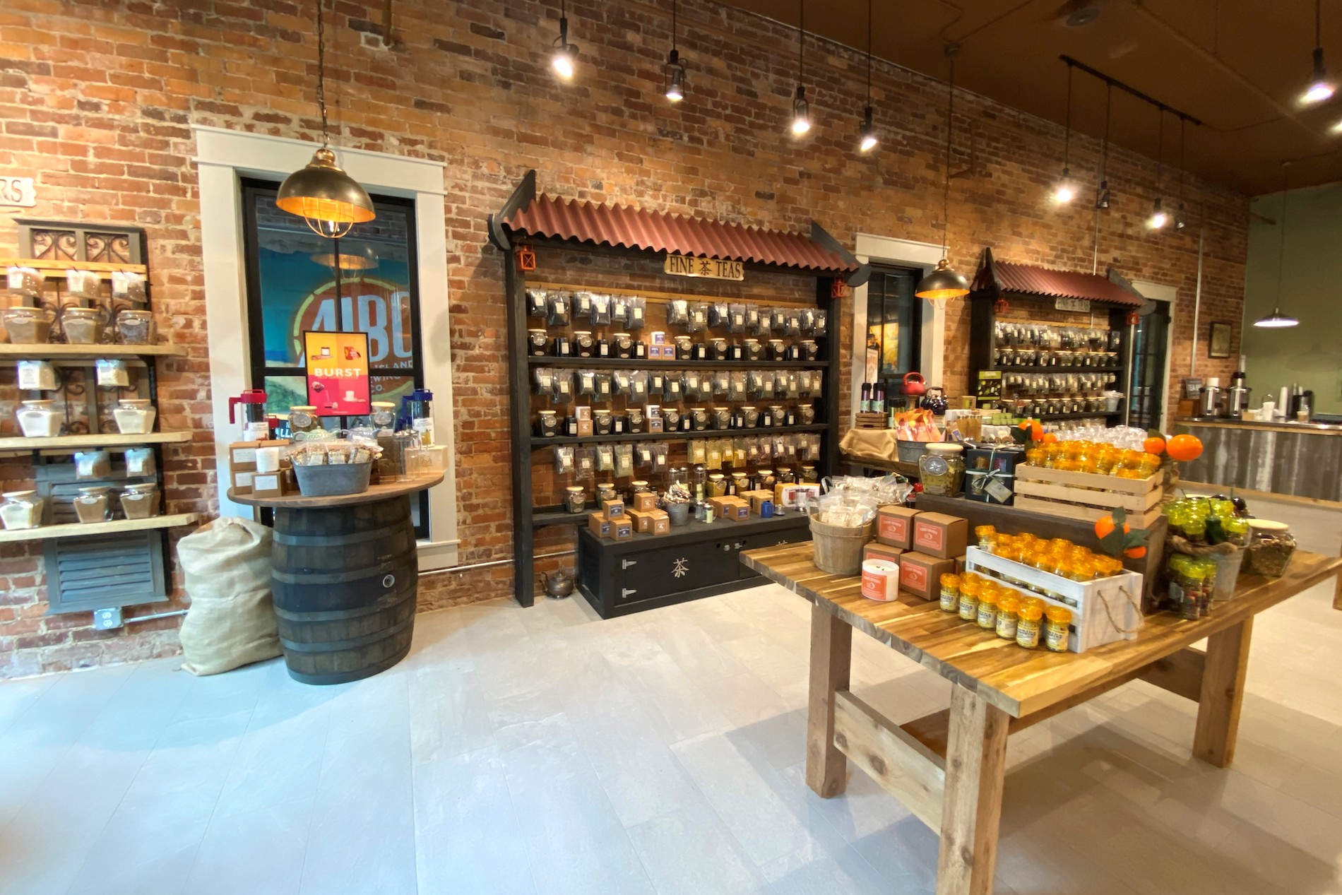 A rustic store interior with brick walls, shelves of tea canisters, baskets, packaged goods, a wooden barrel display, and a table with honey jars and gift items under warm lighting.