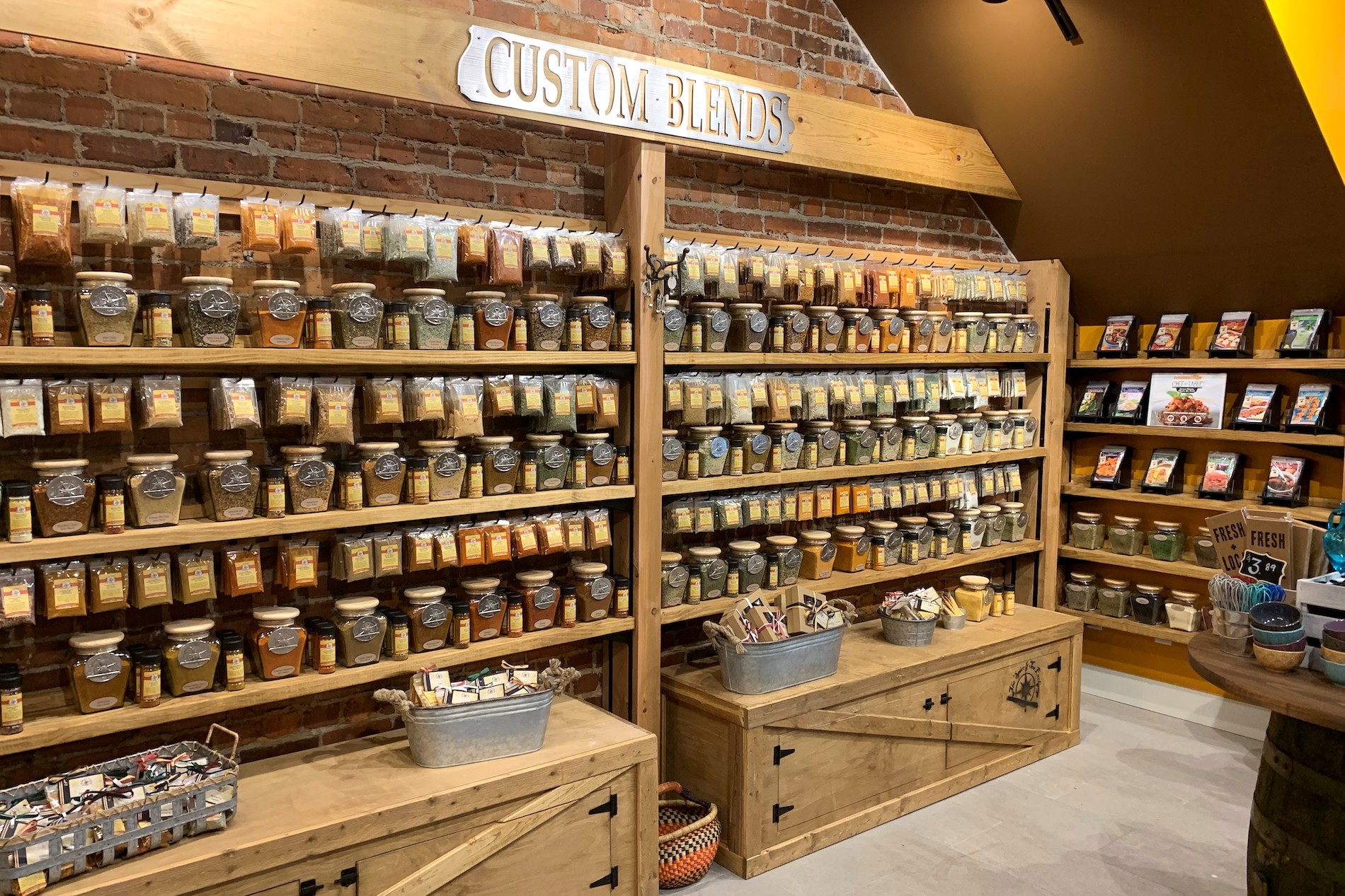 Shelves filled with glass jars of spices and seasonings labeled Custom Blends, with baskets of smaller items and spice packets on wooden cabinets under rustic, warm lighting.