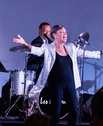 A woman in a silver jacket sings passionately on stage with her arms outstretched, while a drummer plays behind her.