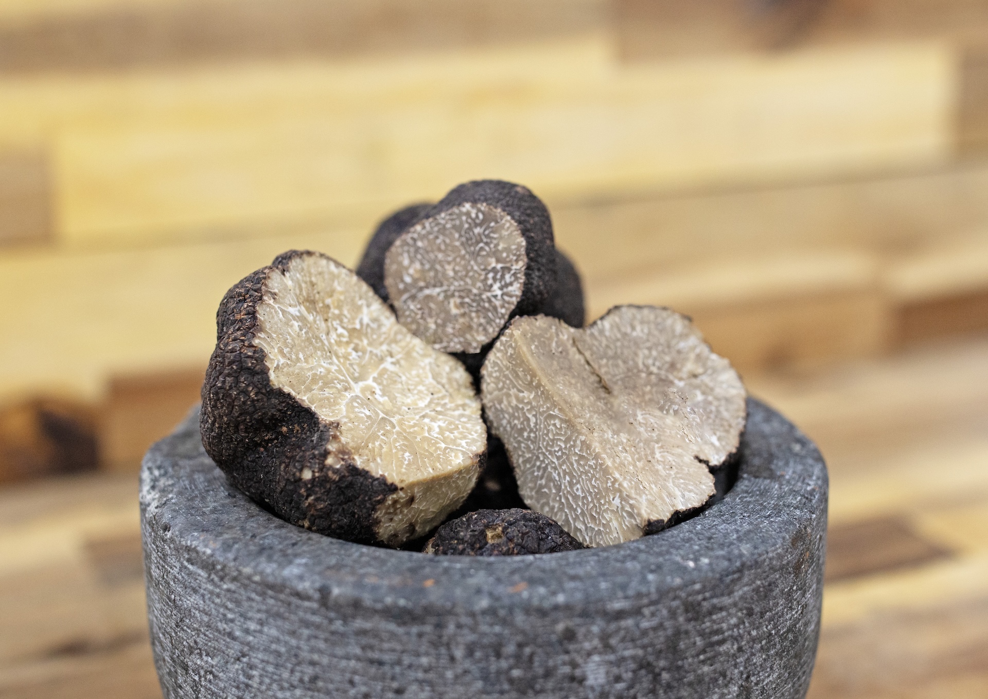 Several black truffles, some sliced open to reveal their marbled interior, sit in a rough, gray stone bowl on a wooden surface with a blurred background.