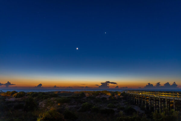 A wooden boardwalk crosses sandy dunes toward a calm ocean at twilight, with the moon and a bright planet visible in a deep blue sky above the orange and yellow glow of the setting or rising sun.