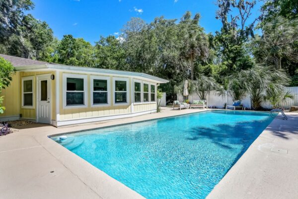 A bright yellow house with large windows sits beside a clear blue swimming pool, surrounded by a concrete deck and lush green trees under a sunny, blue sky.