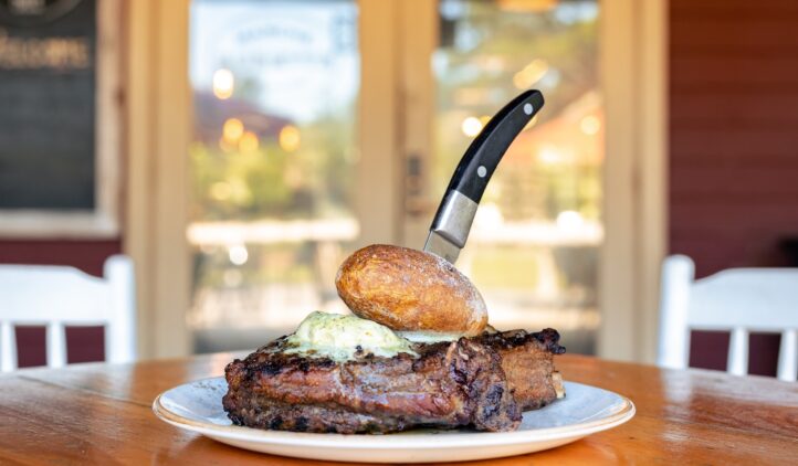 grilled steak topped with herb butter and served with a baked potato at Marche Burette on Amelia Island during the Steak Out event. The dish sits on a wooden table with a blurred restaurant background.