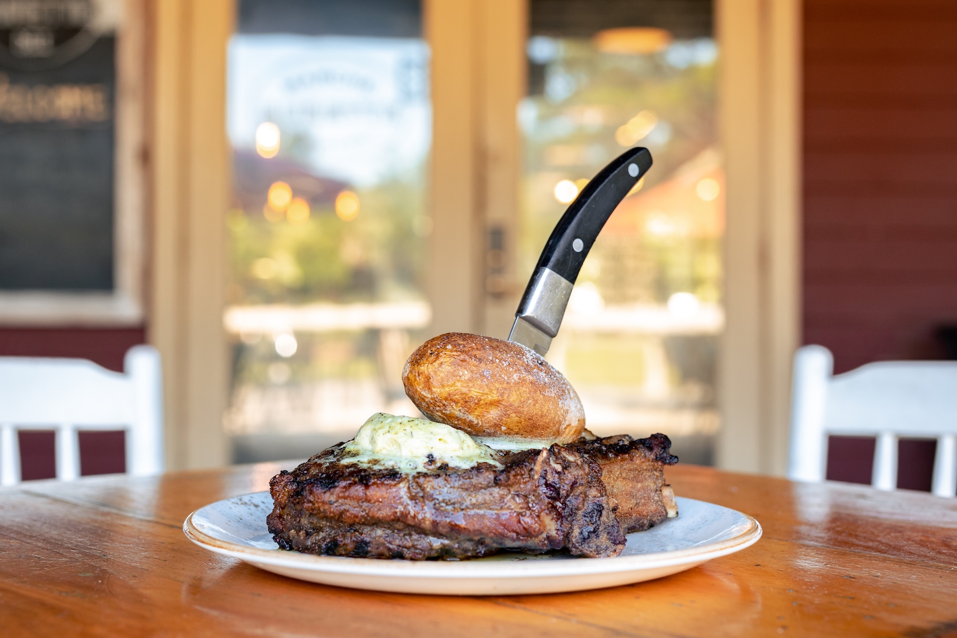 grilled steak topped with herb butter and served with a baked potato at Marche Burette on Amelia Island during the Steak Out event. The dish sits on a wooden table with a blurred restaurant background.