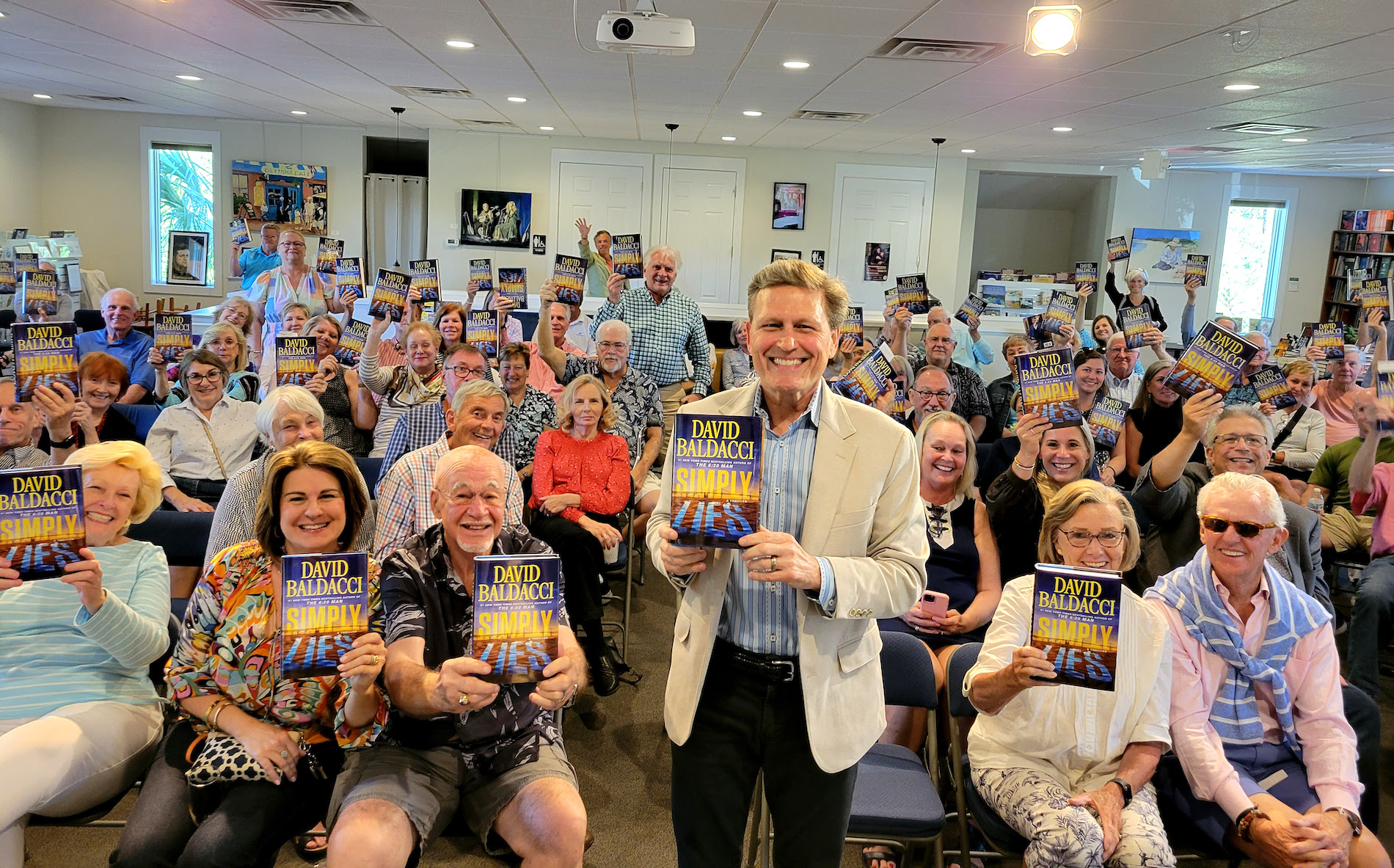 A smiling man in a light suit holds a book titled Simply Lies while posing with a large, cheerful audience, many of whom are also holding up copies of the same book in a brightly lit room.