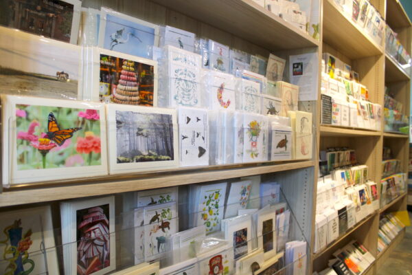 Shelves filled with colorful greeting cards and postcards featuring various designs, such as animals, nature scenes, and artistic illustrations, displayed in a well-lit store.