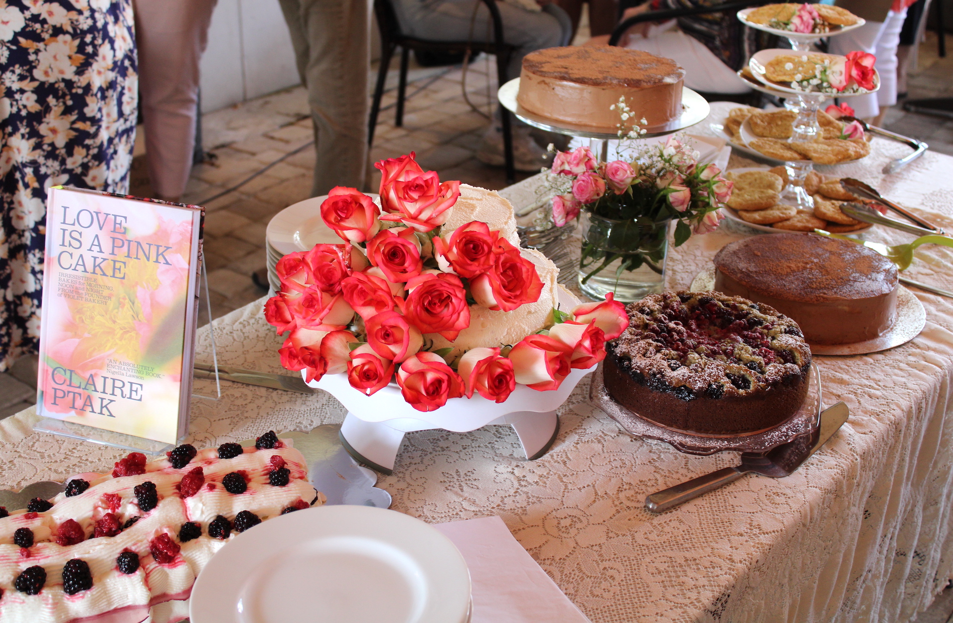 A table covered with a lace cloth displays various cakes, a bouquet of pink roses, and a book titled Love Is a Pink Cake by Claire Ptak. Plates and cutlery are arranged nearby.