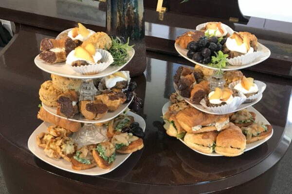 Two tiered serving trays hold assorted pastries, scones, sandwiches, grapes, brownies, and small desserts, set on a dark polished table in a well-lit room.
