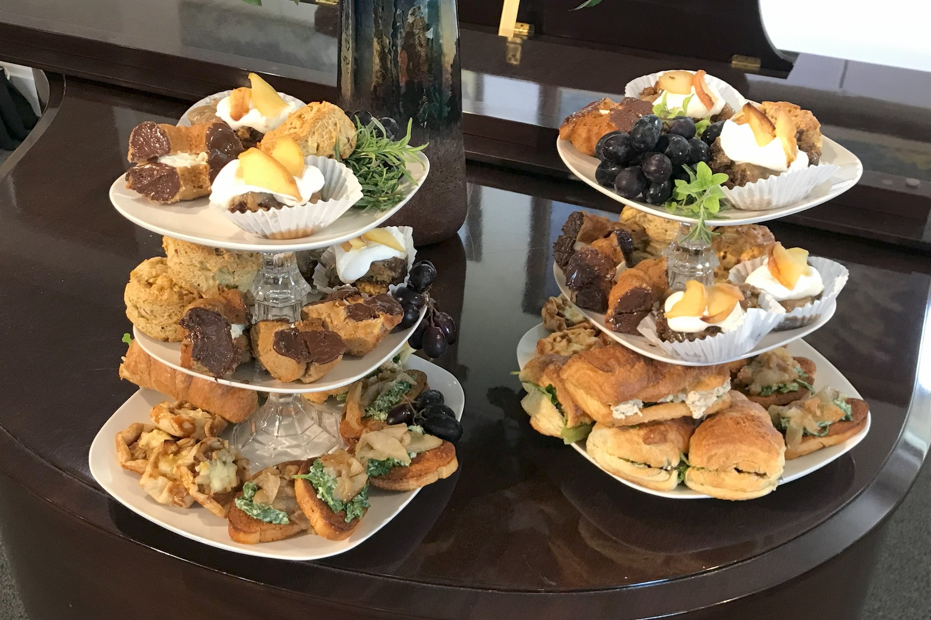 Two tiered serving trays hold assorted pastries, scones, sandwiches, grapes, brownies, and small desserts, set on a dark polished table in a well-lit room.