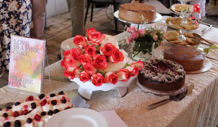 A table covered with a lace cloth displays various cakes, cookies, a plate of blackberries, a bouquet of pink roses, and a book titled LOVE IS A PINK CAKE by Claire Ptak.