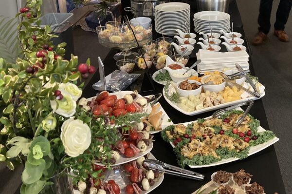 A buffet table with various appetizers, cheeses, meats, garnished salads, desserts, plates, napkins, and utensils, alongside a floral arrangement. A person’s legs are visible in the background.