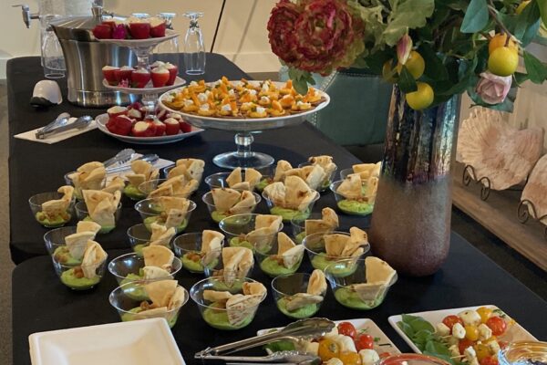 Elegant buffet table with small dishes of pita and dip, stuffed strawberries, bite-sized appetizers on a tray, a flower vase, and a cherry tomato salad in the foreground.