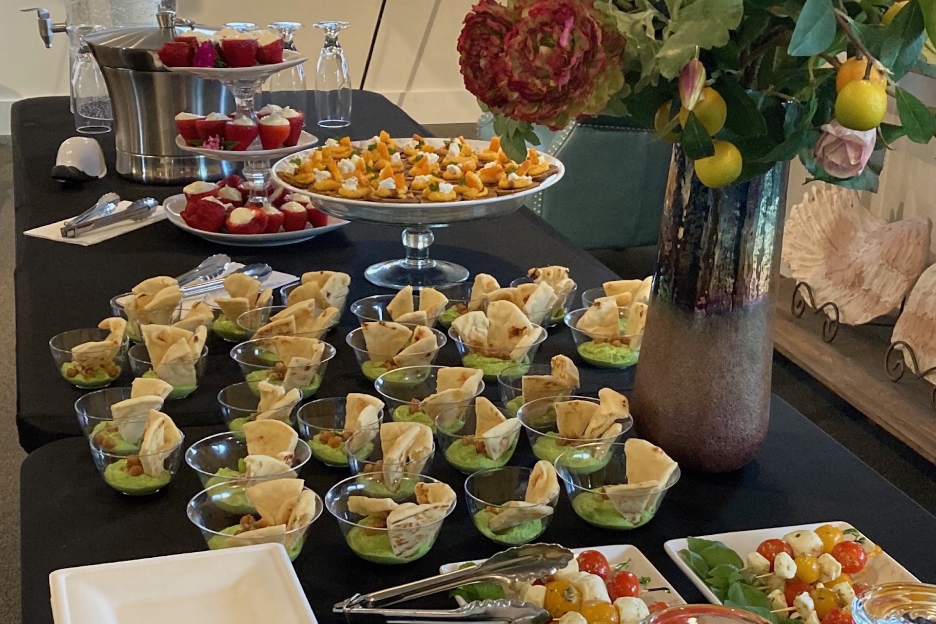 Elegant buffet table with small dishes of pita and dip, stuffed strawberries, bite-sized appetizers on a tray, a flower vase, and a cherry tomato salad in the foreground.