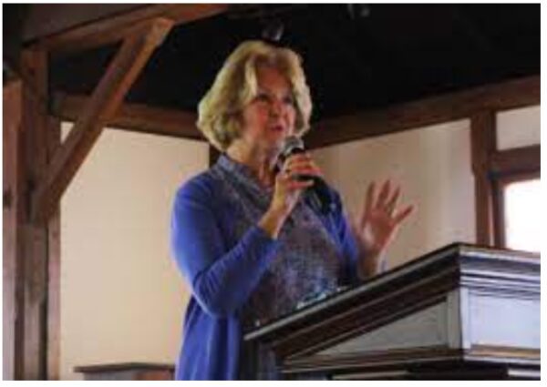 A woman with blonde hair wearing a blue sweater speaks into a microphone while standing at a wooden podium in a room with wooden beams and natural light.