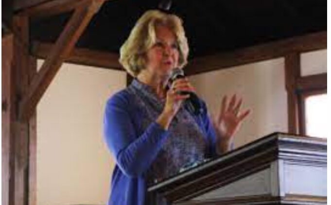 A woman with blonde hair wearing a blue sweater speaks into a microphone while standing at a wooden podium in a room with wooden beams and natural light.