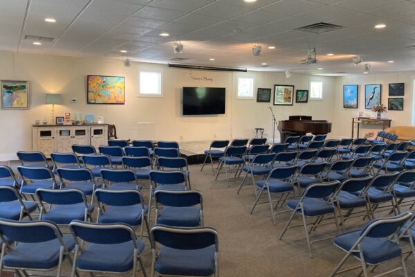 A small event room with rows of blue folding chairs facing a stage with a TV, piano, and artwork on the walls. The room has white walls, ceiling lights, and windows letting in natural light.